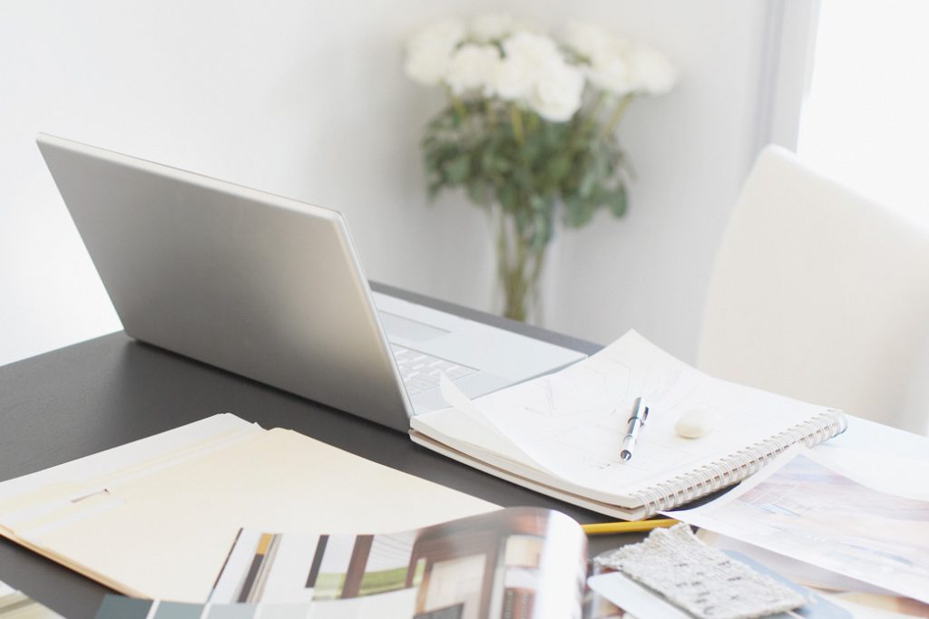 Desk with notebook and pieces of paper, with open laptop and white chair.